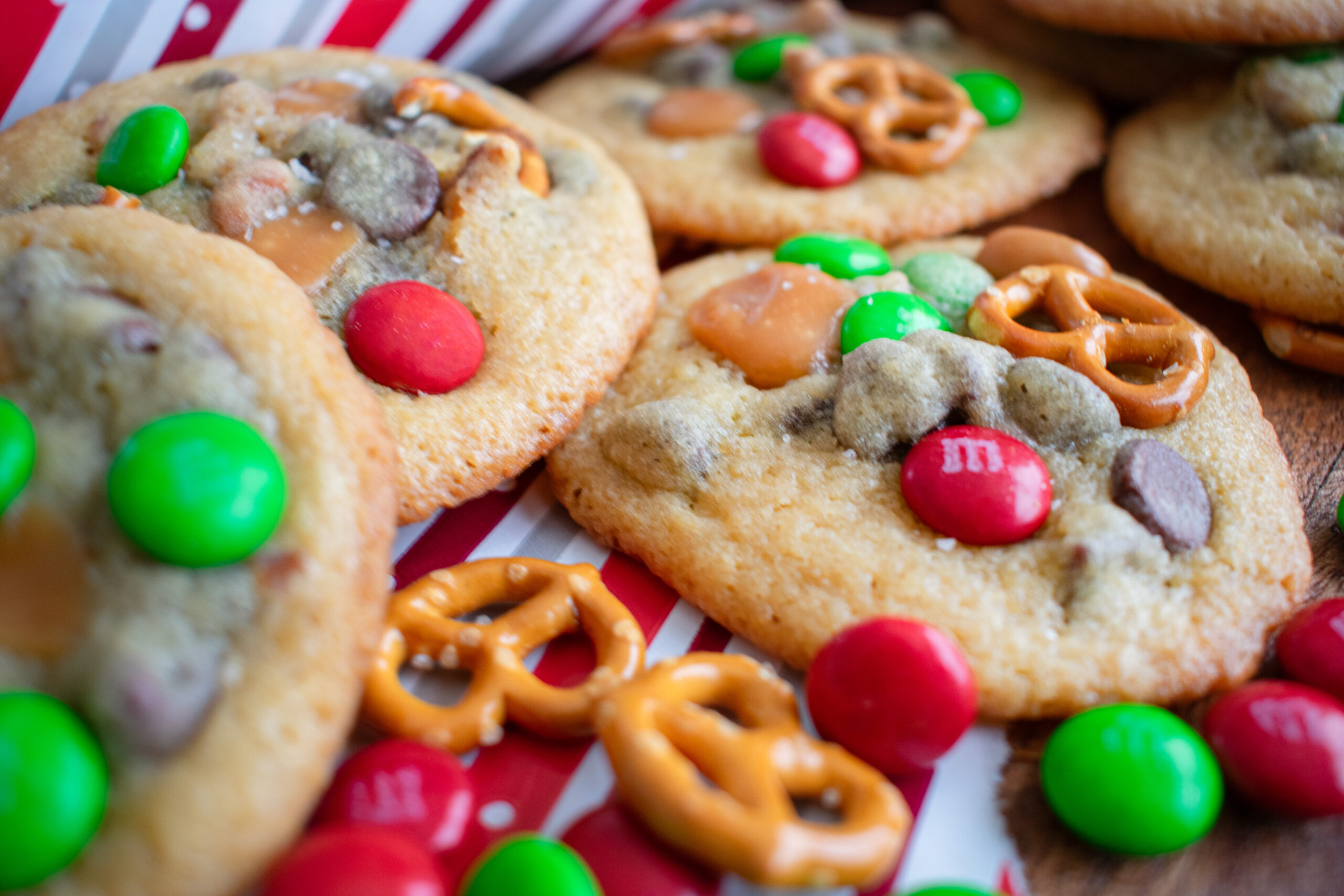 Festive Kitchen Sink Cookies loaded with chocolate chips, pretzels, holiday candy, and more on a red and white stripe, perfect for holiday parties and cookie swaps.