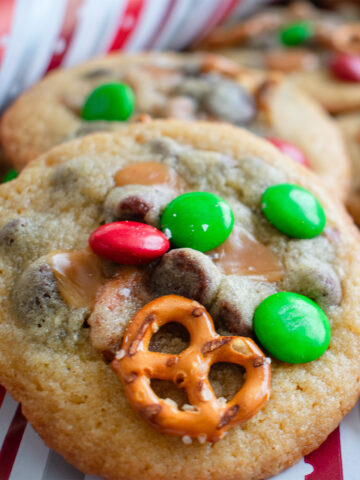 Festive Kitchen Sink Cookies loaded with chocolate chips, pretzels, holiday candy, and more on a red and white stripe, perfect for holiday parties and cookie swaps.