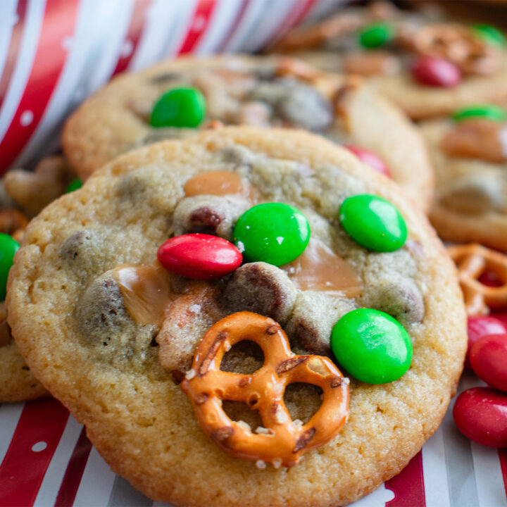 Festive Kitchen Sink Cookies loaded with chocolate chips, pretzels, holiday candy, and more on a red and white stripe, perfect for holiday parties and cookie swaps.