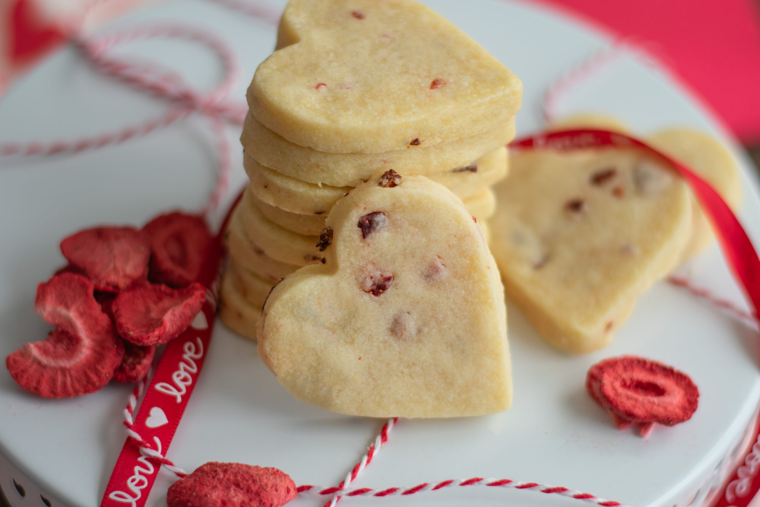 Stacked strawberry flecked shortbread cookies with strawberries scattered nearby and ribbons, showcasing their buttery texture and fruity accents