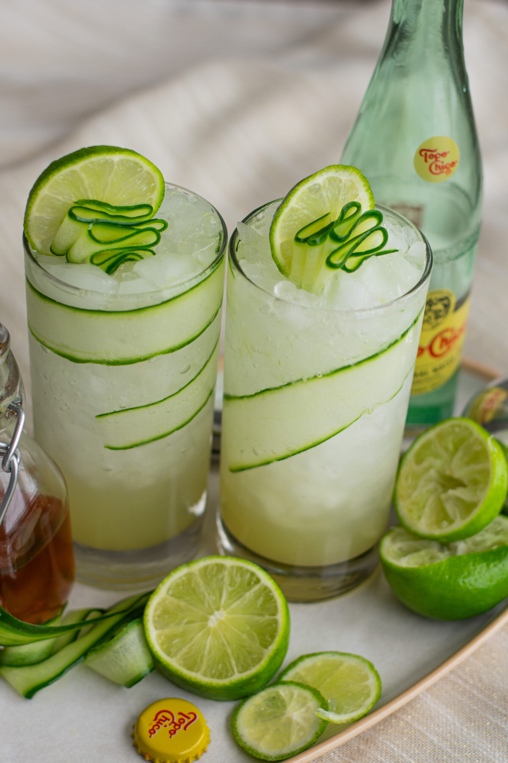 Cucumber Gimlet in a tall glass with cucumber ribbons, lime wedges, a water bottle, and icy beverage for a refreshing mocktail experience.