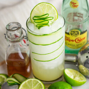 Cucumber Gimlet in a tall glass with cucumber ribbons, lime wedges, a water bottle, and icy beverage for a refreshing mocktail experience.