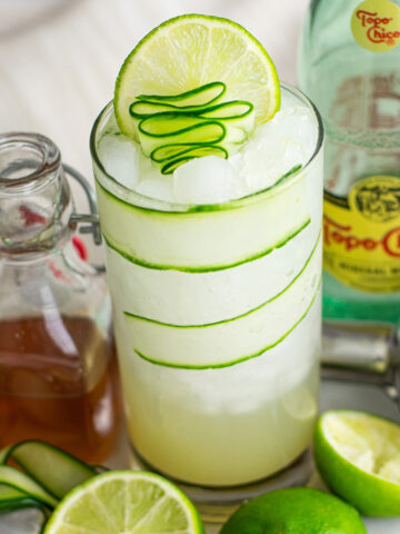 Cucumber Gimlet in a tall glass with cucumber ribbons, lime wedges, a water bottle, and icy beverage for a refreshing mocktail experience.