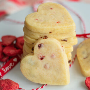 Stacked strawberry flecked shortbread cookies with strawberries scattered nearby, showcasing their buttery texture and fruity accents