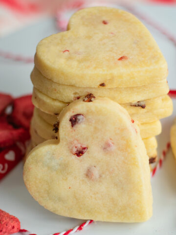 Stacked strawberry flecked shortbread cookies with strawberries scattered nearby, showcasing their buttery texture and fruity accents