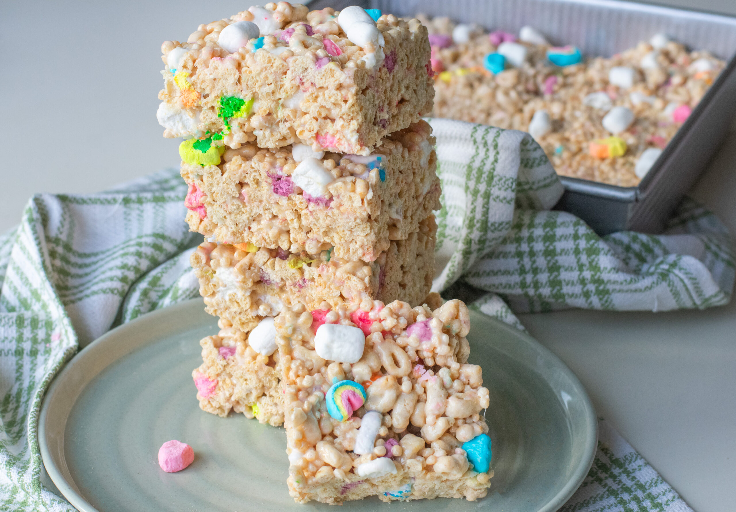 Lucky Charms Marshmallow Treat Bars Stacked High near a tray of Rice Krispie Treats