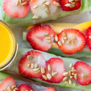 Fresh spring rolls filled with colorful vegetables on a white plate, served with a small bowl of ginger dipping sauce on the side.