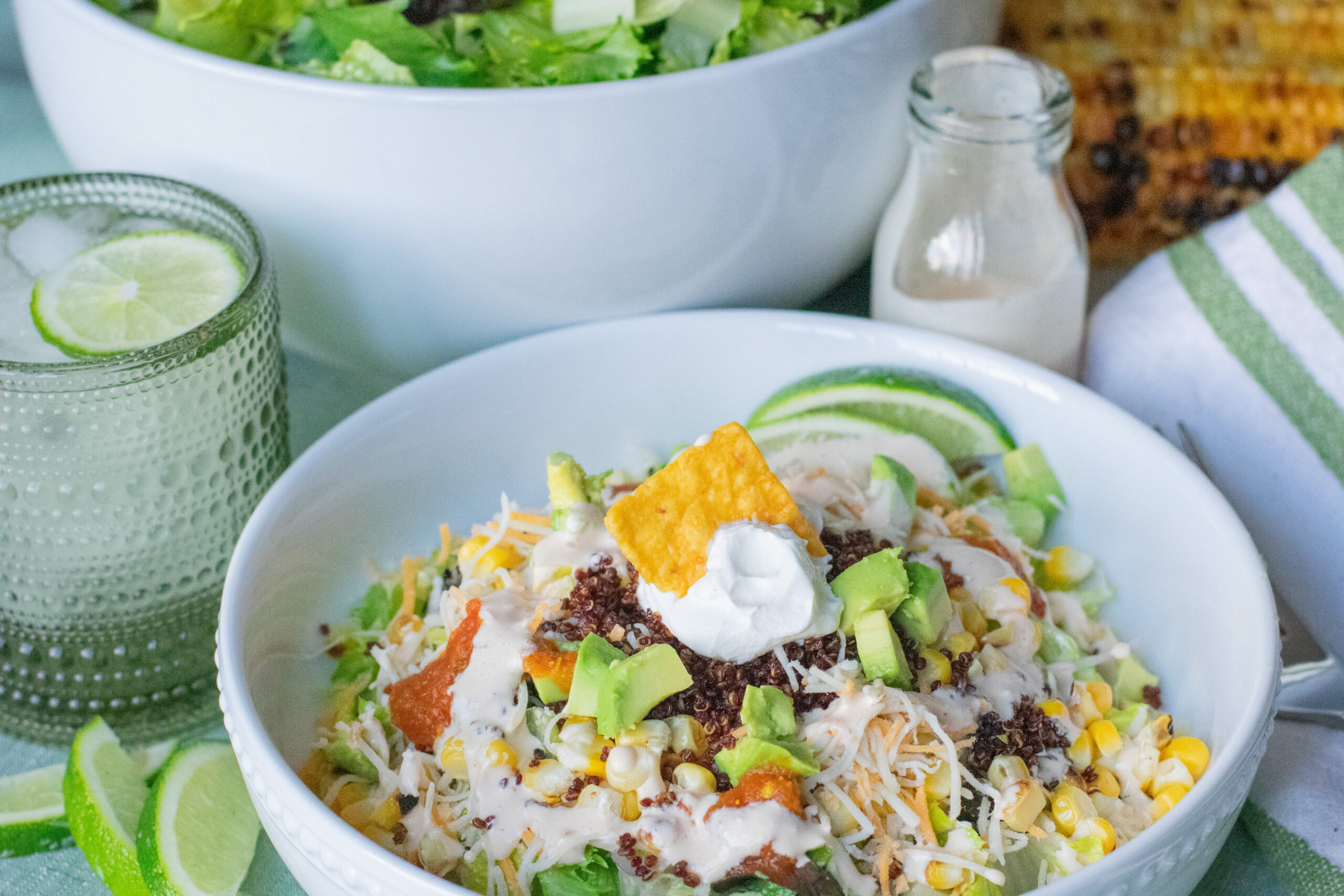 Vegetarian taco salad served in a bowl next to a glass of creamy spicy dressing, fresh corn on the cob, and a second salad bowl in the background.