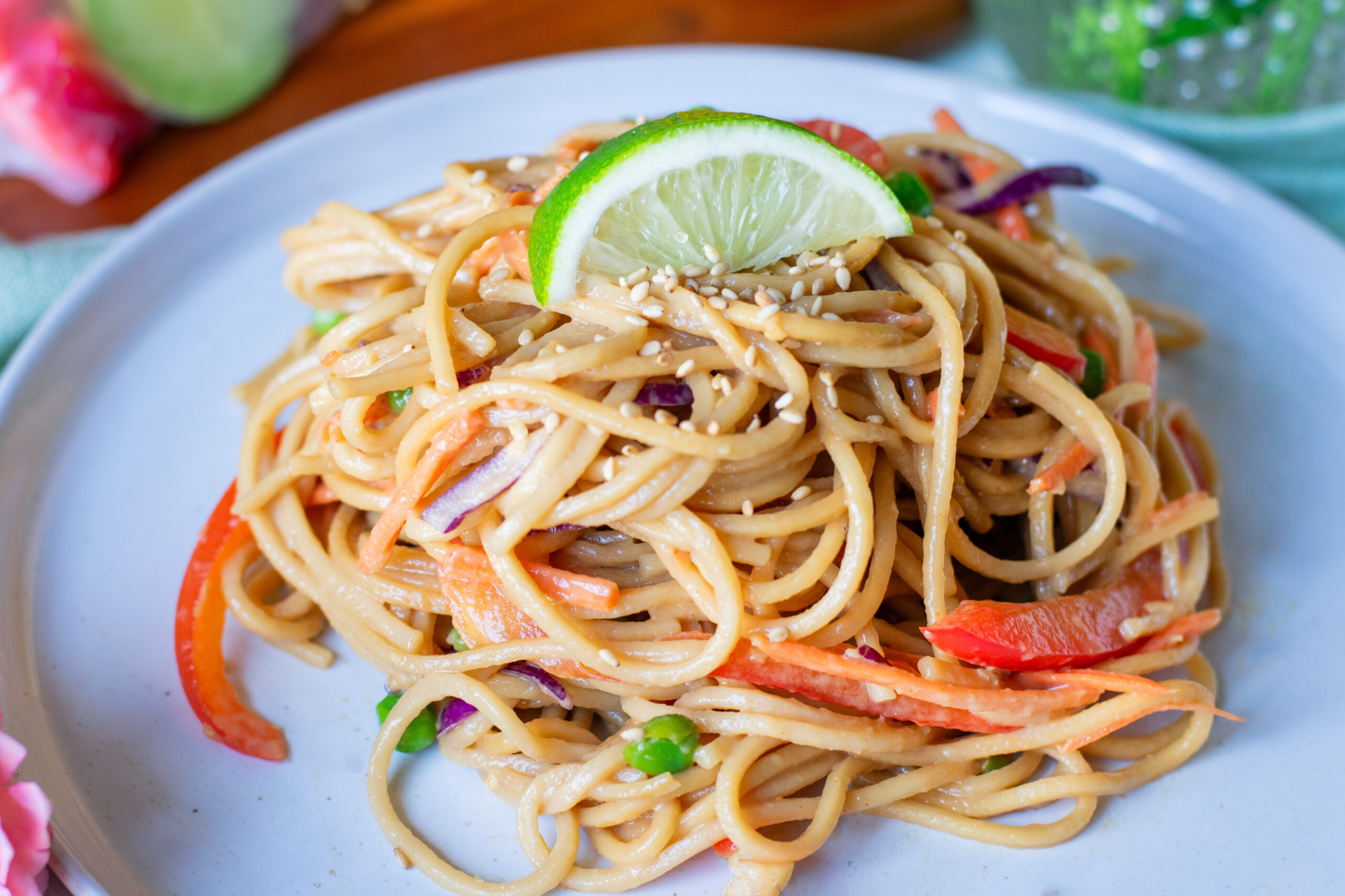 Close-up of cold vegetable lo mein topped with sesame seeds and a lime slice, with fresh flowers nearby