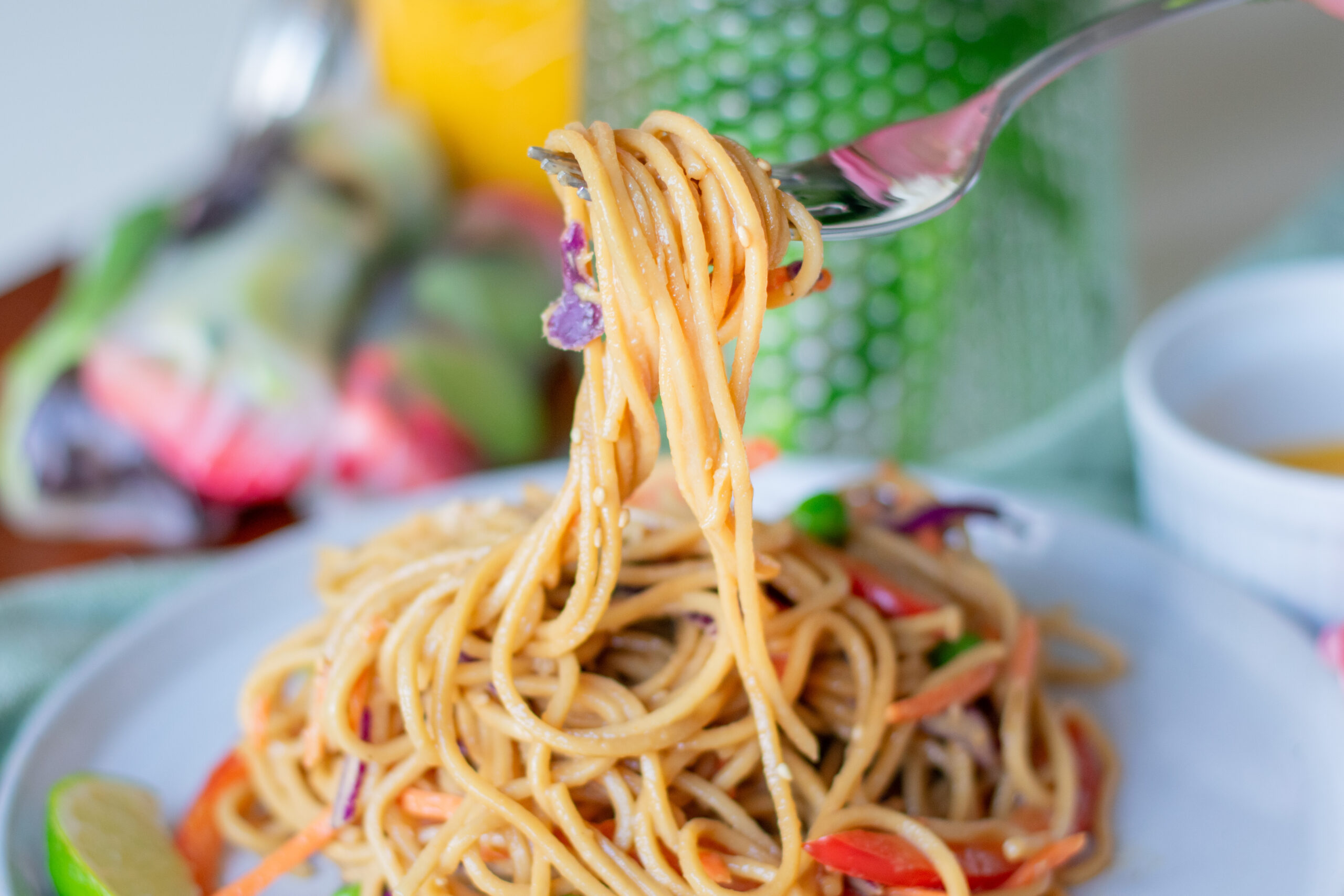 Fork lifting cold vegetable lo mein noodles above a bowl, with fresh flowers nearby.