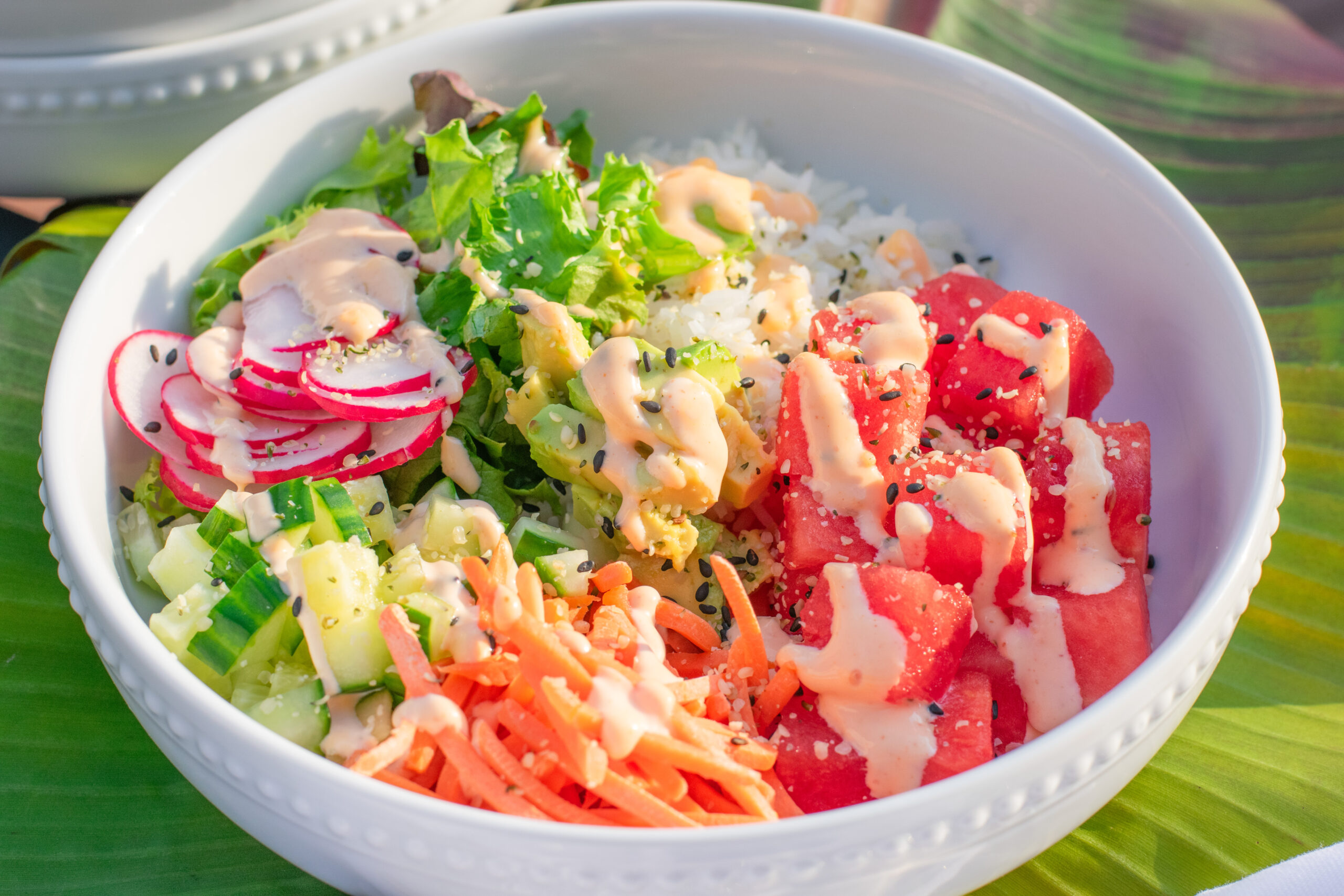Watermelon poke bowl served over a tropical banana leaf, with white rice, avocado, cucumber, radish, carrots, and a spicy mayo drizzle.