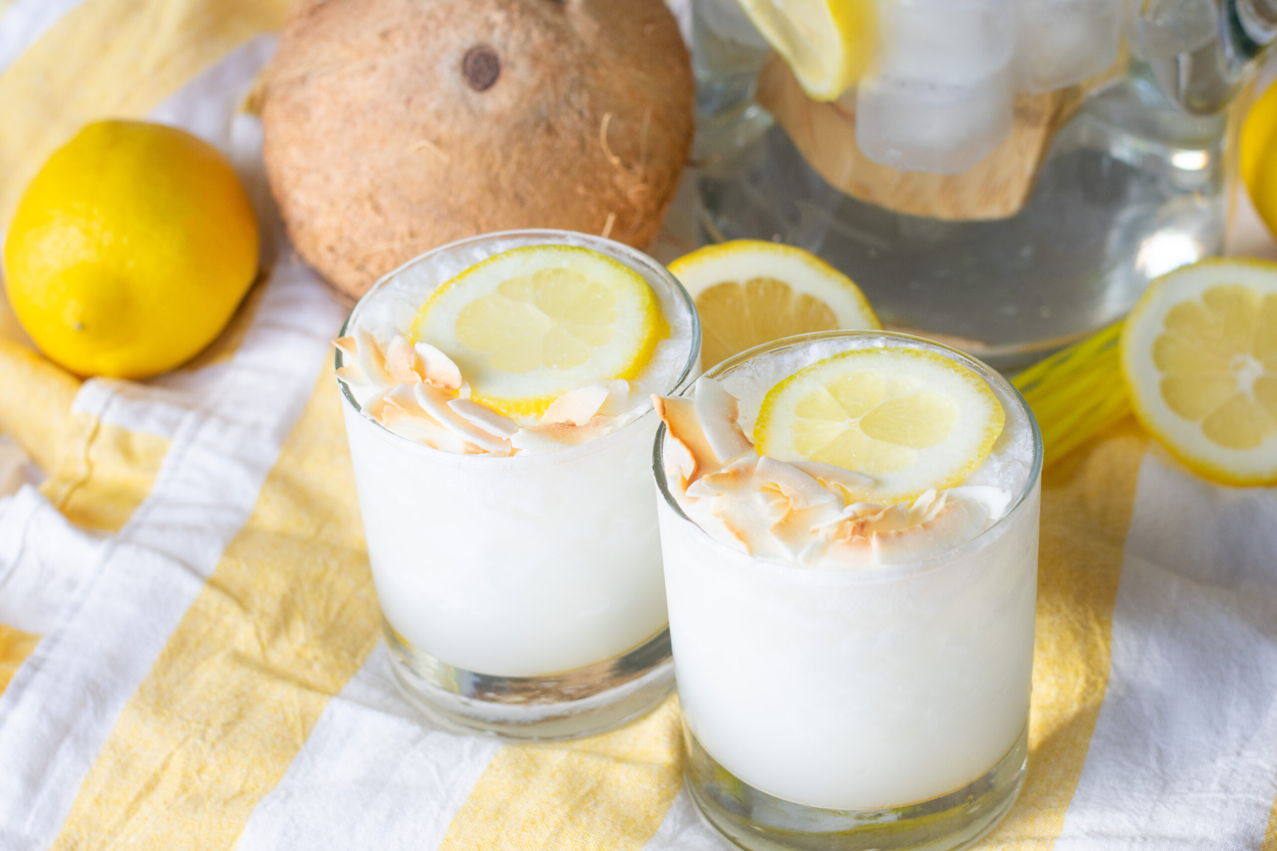 Two glasses of creamy Lemoncoco lemonade beside a fresh coconut and a pitcher of the drink on a wooden surface