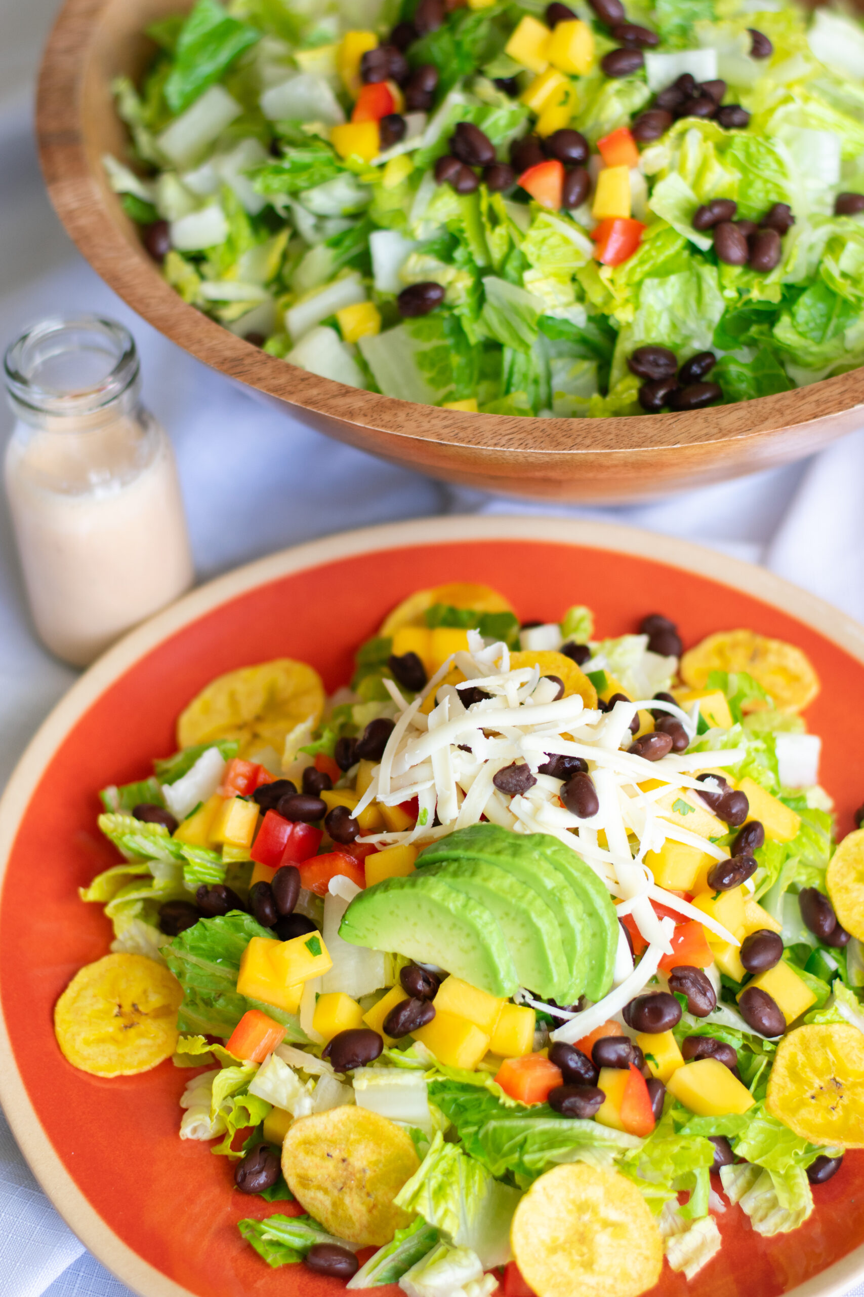 Colorful Mango Fiesta Salad on a red plate with avocado, black beans, cheese, mango salsa, and creamy ranch dressing, served with plantain chips.