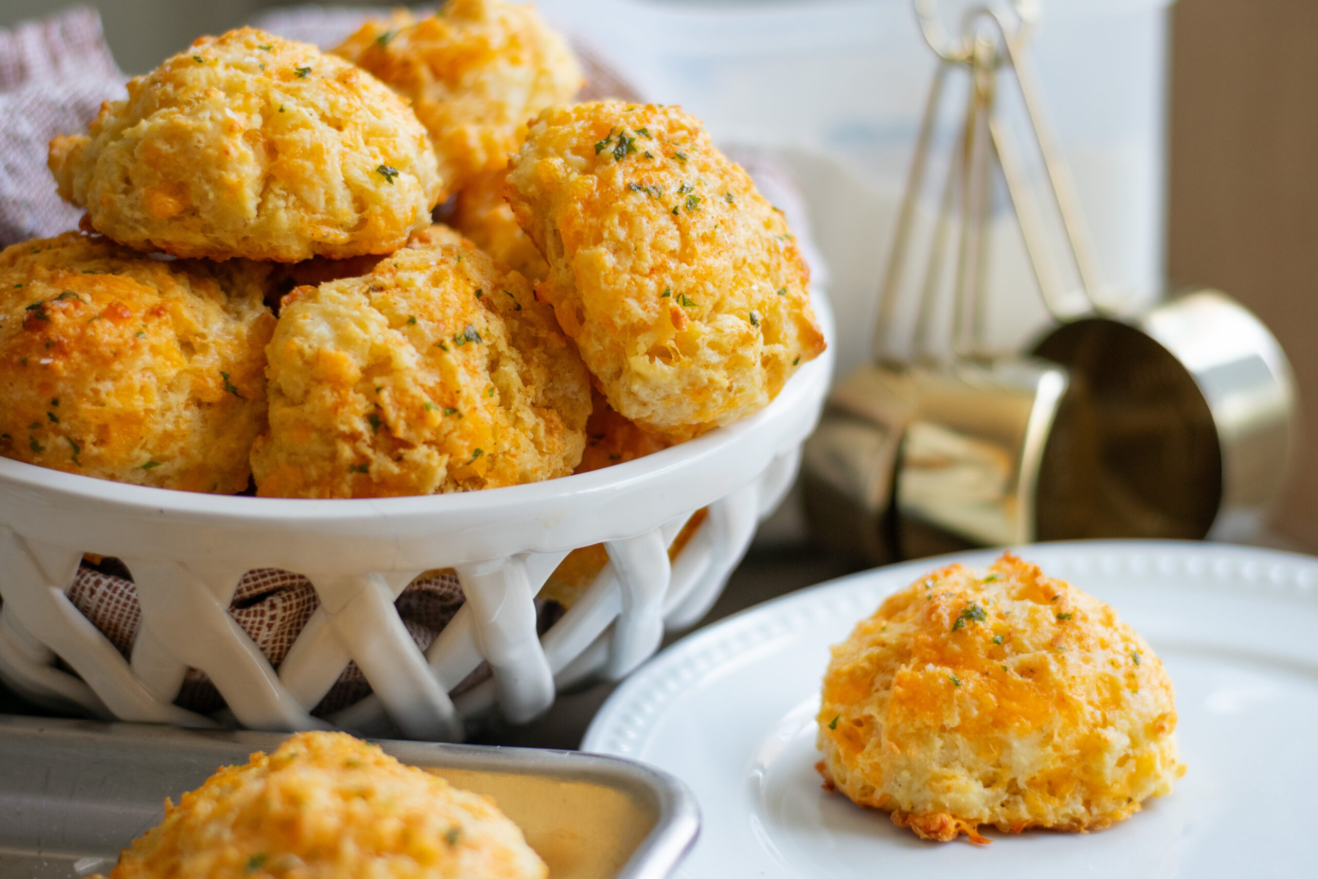 Large close-up of a golden homemade buttermilk cheddar biscuit on a plate, with a pile of cheesy garlic biscuits behind it, brushed with melted garlic butter and garnished with parsley.