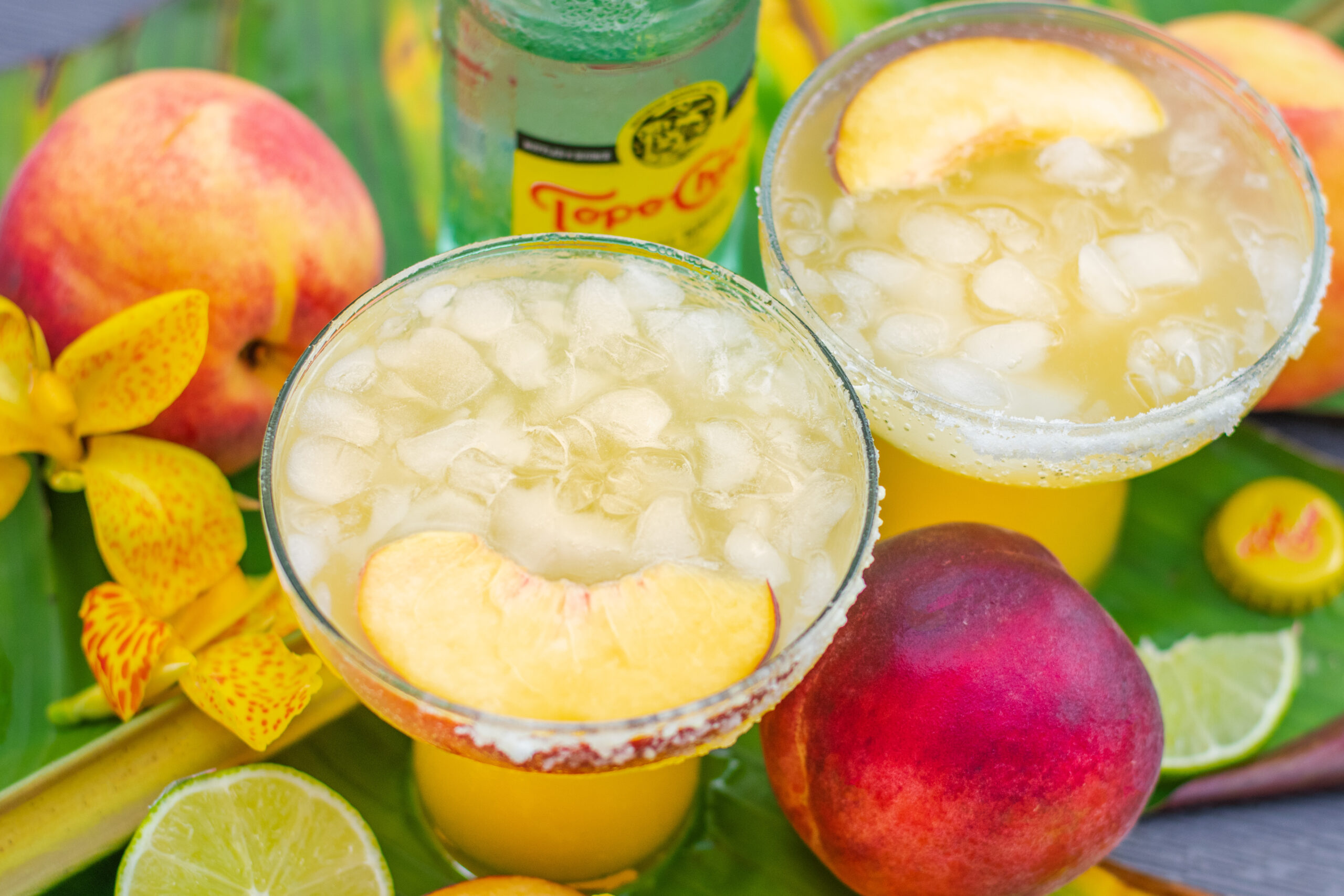 Peach margarita ingredients scene with ripe peaches, flowers, and Topo Chico sparkling mineral water bottle and two glasses.