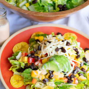 Colorful mango fiesta salad on a red plate with avocado, black beans, shredded cheese, mango salsa, and creamy ranch dressing, served with plantain chips and a large wooden bowl of salad in the background.