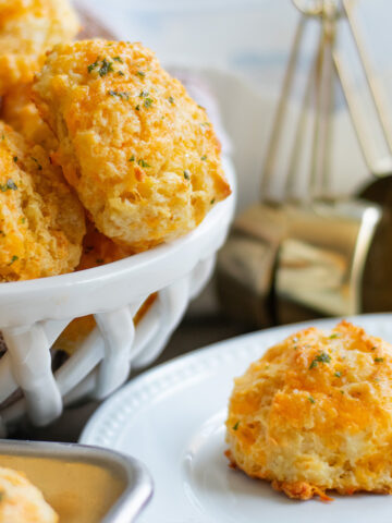 Golden homemade buttermilk cheddar biscuit on a white plate with a pile of cheesy garlic biscuits in the background, topped with fresh parsley and brushed with melted garlic butter.