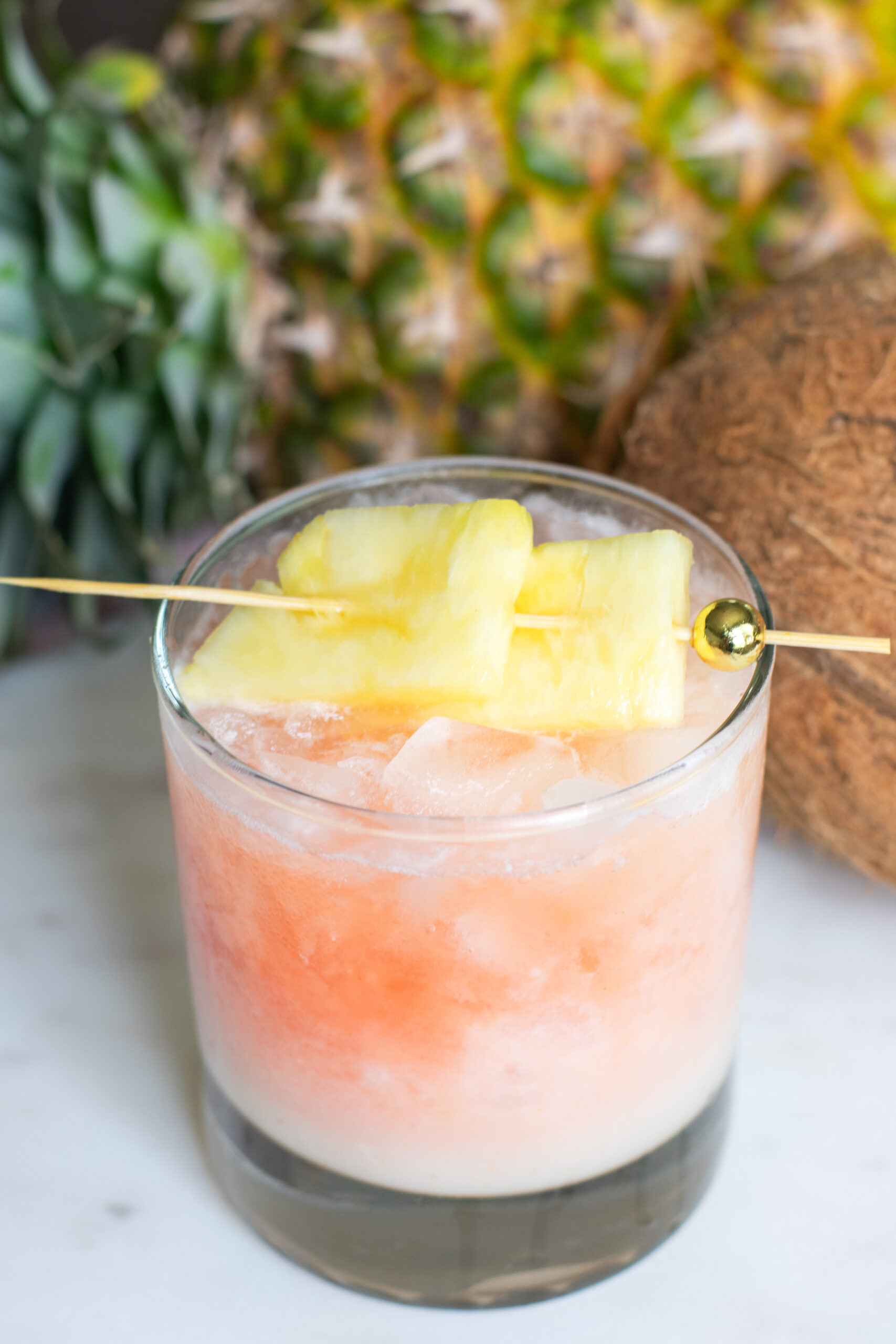 Guava Sunrise Colada in a glass styled with pineapple fronds and whole coconut