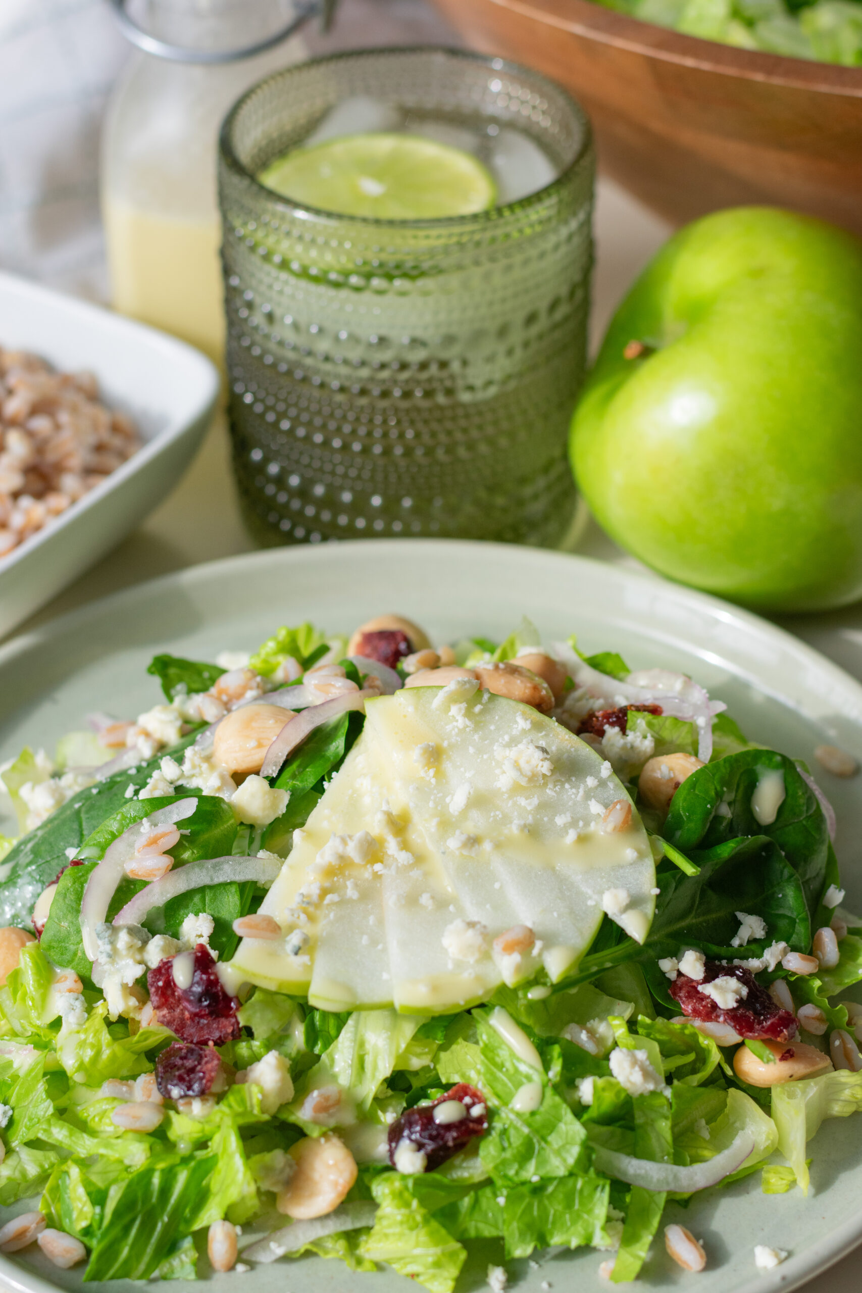 Green apple farro salad with gorgonzola and cranberries shown with cooked farro, apple vinaigrette, fresh green apple, and mixed greens on a table