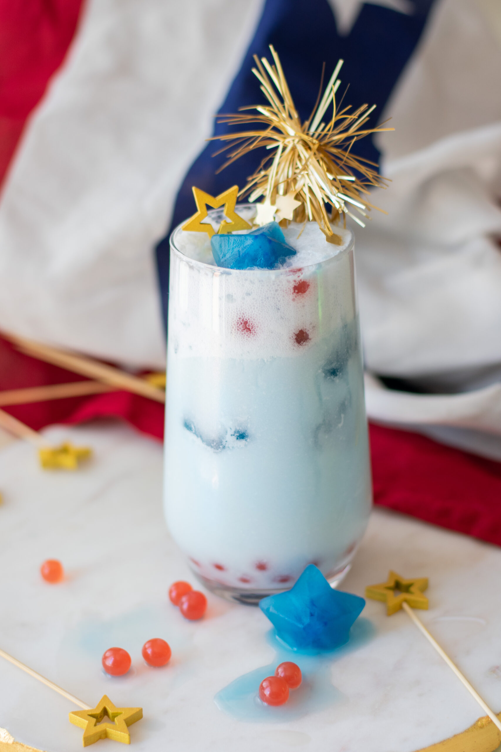 Patriotic red white and blue drink with strawberry popping boba, coconut milk, sparkling soda, and blue star ice cubes.